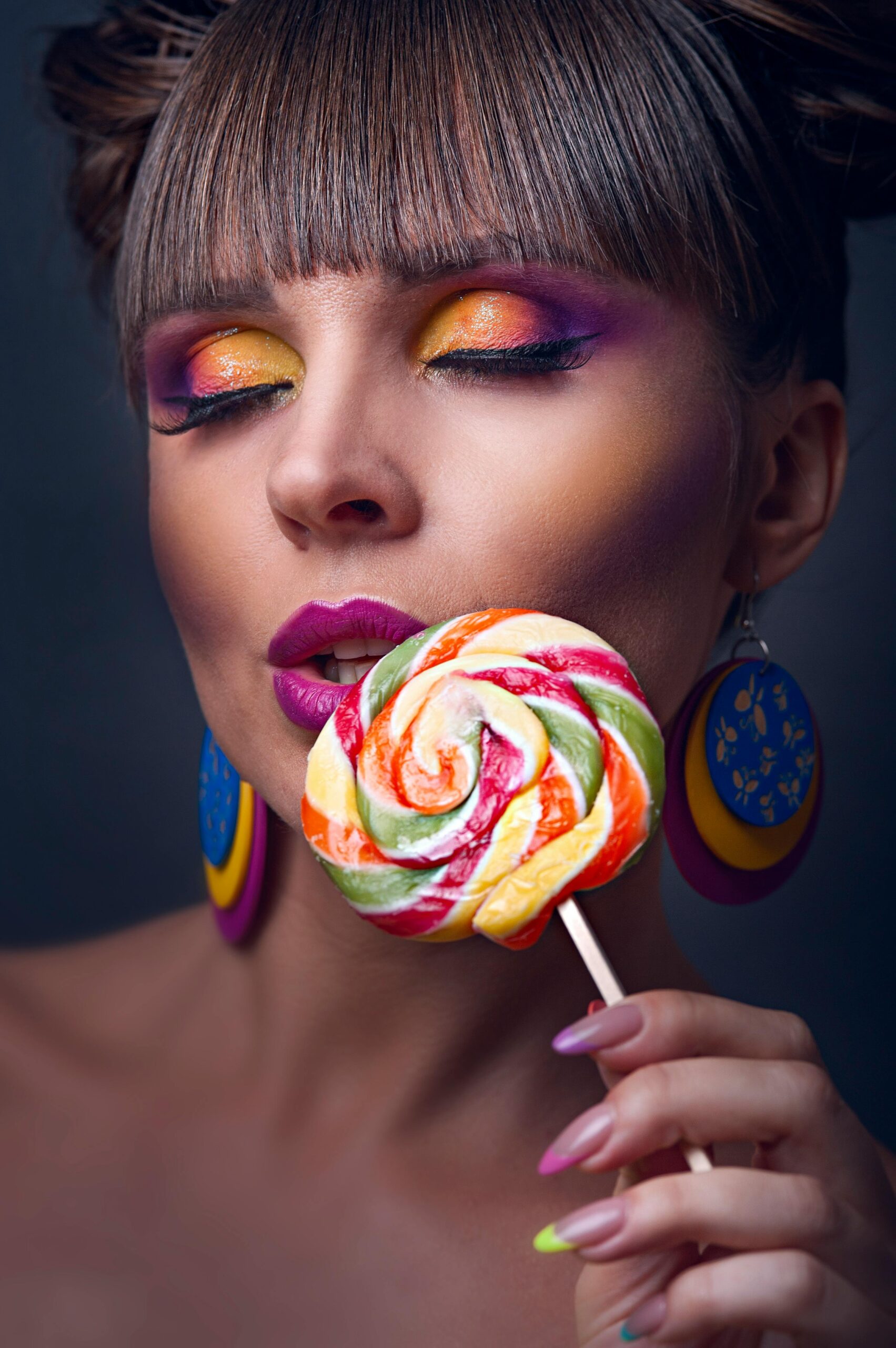 Close-up portrait of a woman with bold colorful makeup and a lollipop.
